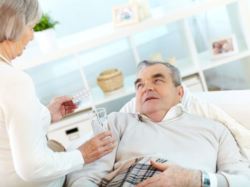 Caregiver handing medication and water to a senior man resting on the couch, part of recognizing medication side effects through close daily attention.