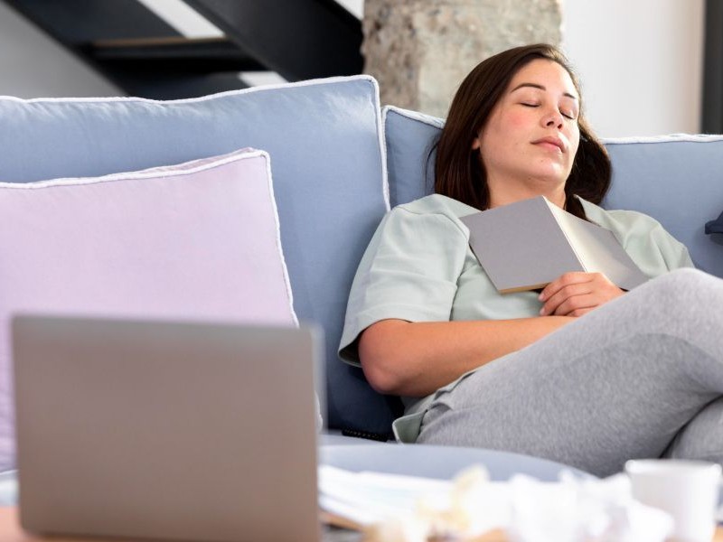 Why Preventing Caregiver Exhaustion Starts With Small Breaks Exhausted woman napping on the couch with a book and laptop nearby, showing why preventing caregiver exhaustion starts with allowing real rest.