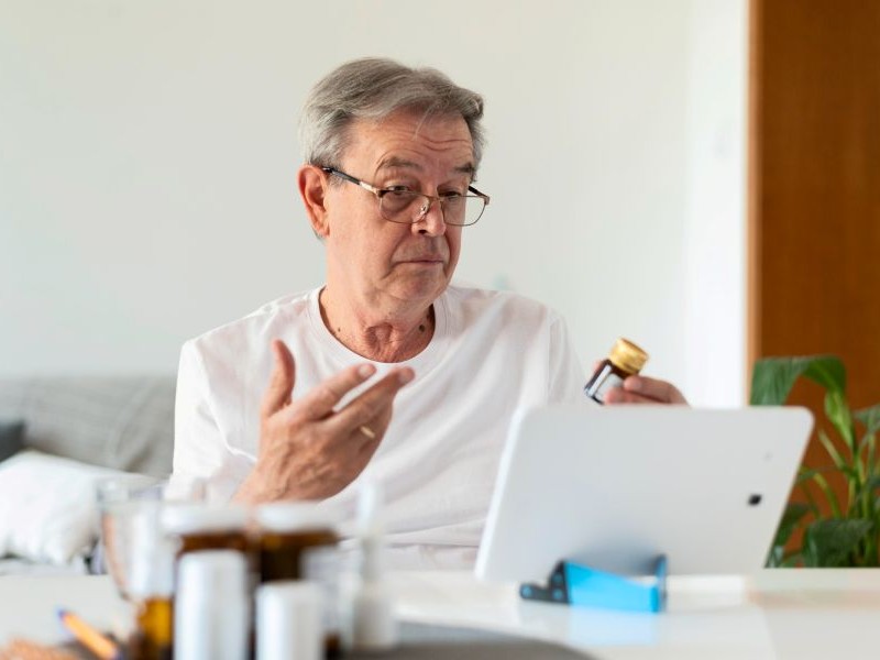 Recognizing Medication Side Effects in Seniors Senior man checking a pill bottle during a video call, an example of recognizing medication side effects before they lead to bigger concerns.